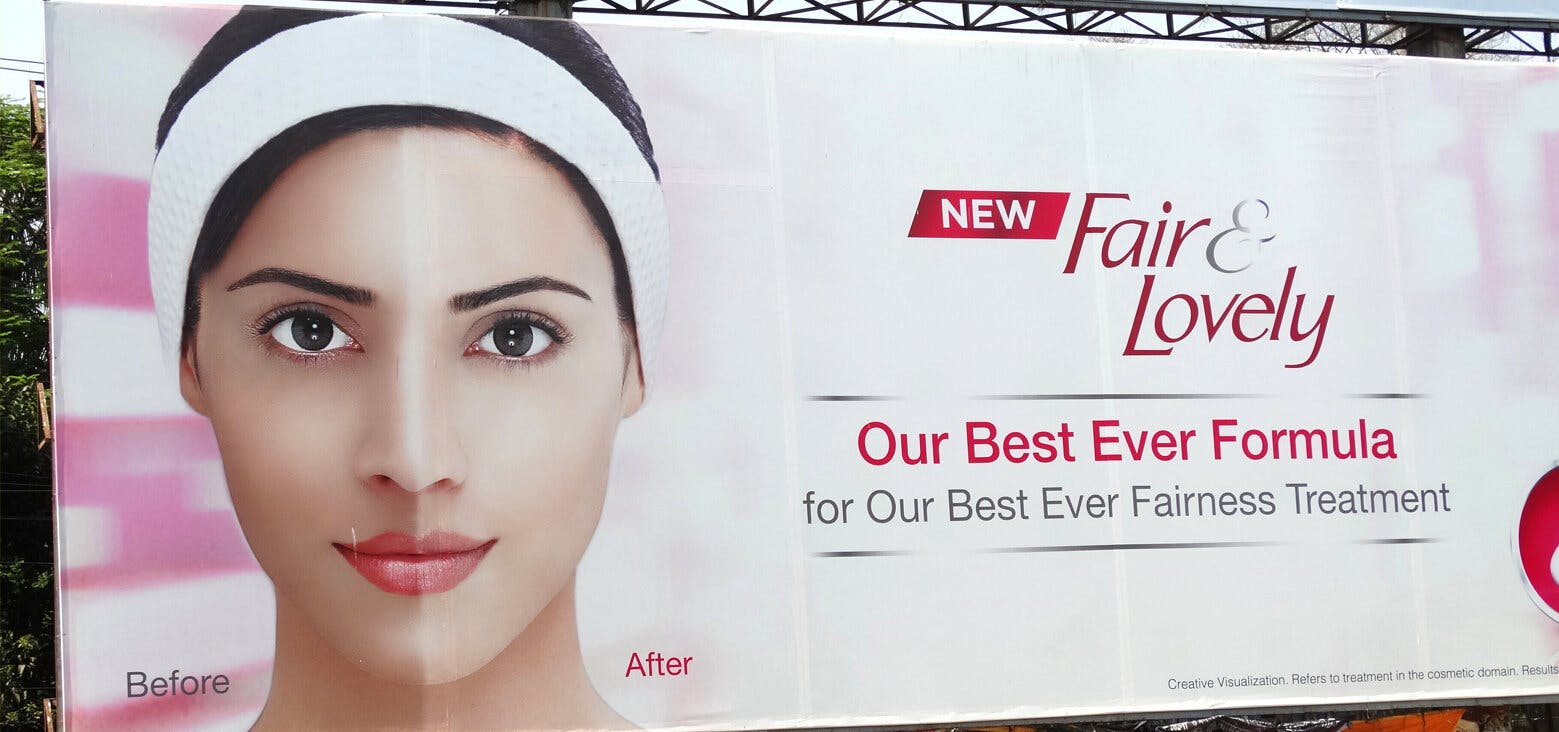 A pink billboard ad for the skin bleaching brand 'Fair & Lovely' in India, where a woman is pictured with half her face darker than the other half, accompanied by the text 'before' and 'after. Next to this is the text 'New / Fair & Lovely / Our Best Ever Formula / for Our Best Ever Fairness Treatment'