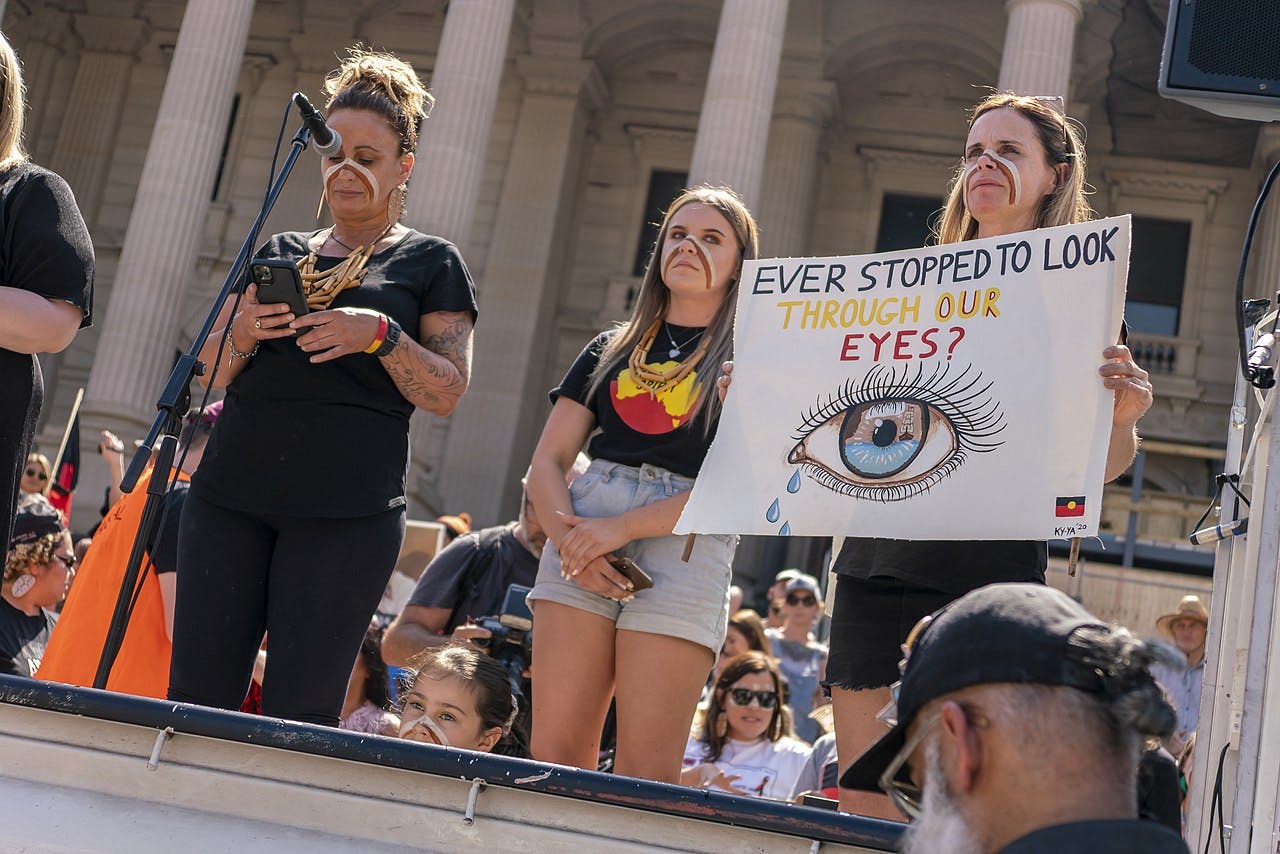 Three people standing on the stage at the 2020 Invasion Day rally in Naarm/Melbourne. The protestor on the right is holding up a sign that reads "Ever stopped to look through our eyes?"
