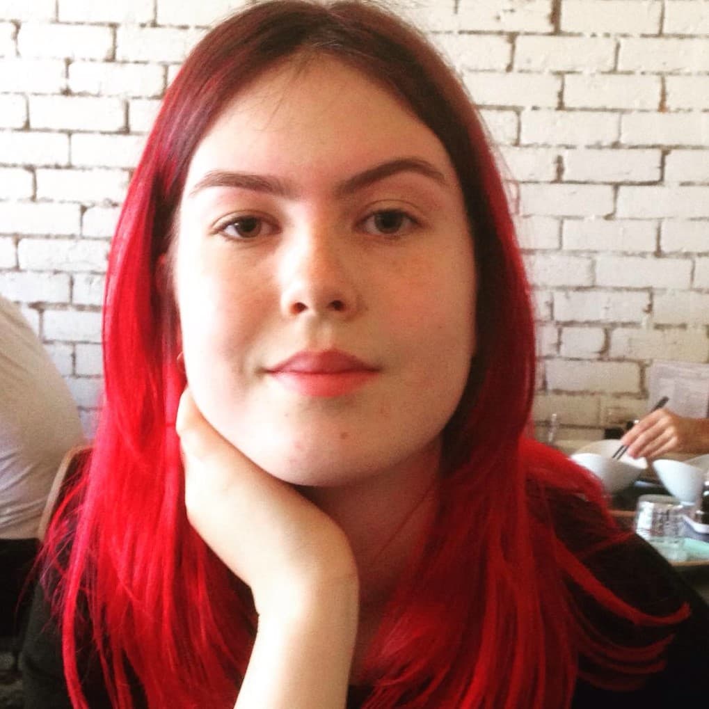 Alicia looking at the camera with her hand to her neck. She has red hair.
