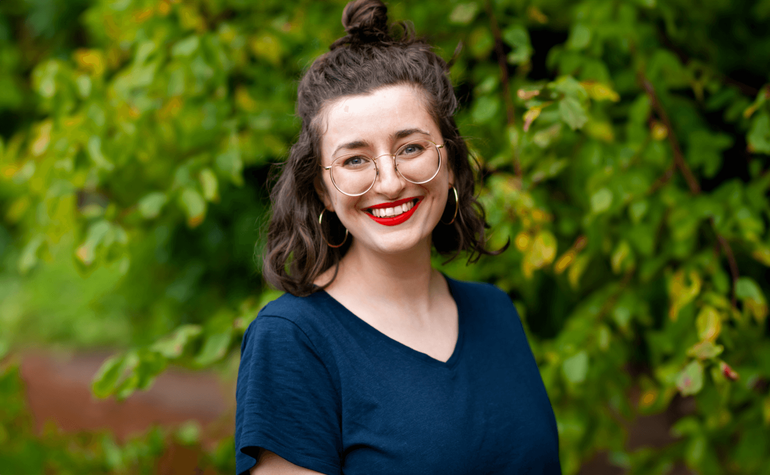 A headshot of Alice Boyle. She wears a navy top, glasses and hoop earings, and is standing before trees.