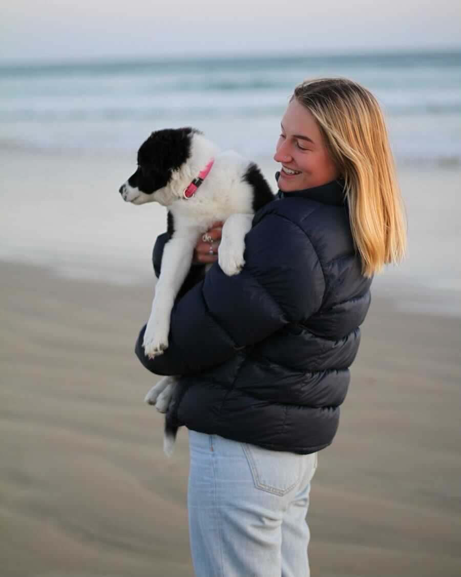 An image of Lily Mooney at the beach holding a dog. She wears a black puffer jacket and blue jeans.