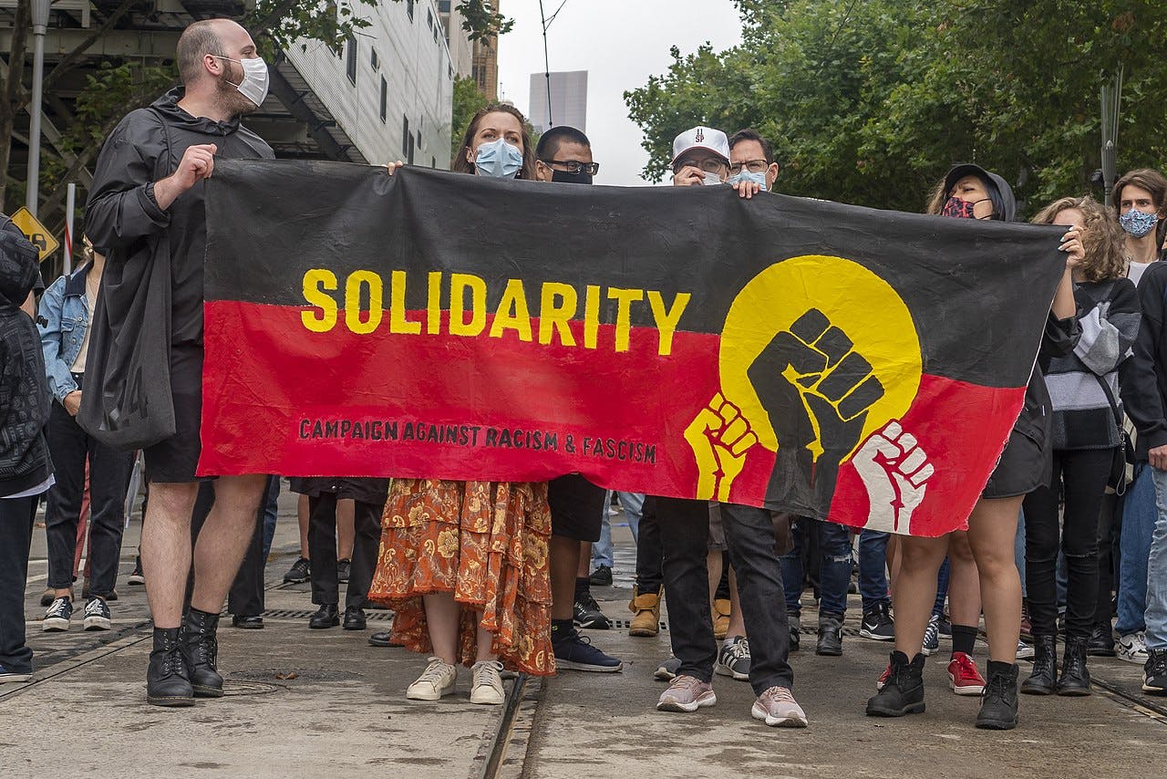 Protestors at the 2021 Invasion Day rally in Naarm/Melbourne holding up a large banner in the colours of the Aboriginal flag that reads "Solidarity / Campaign against racism & facism".